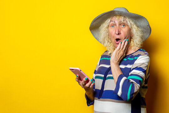 Surprised shocked old woman in a hat holds a phone on a yellow background.