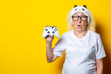 Surprised blonde old woman with joystick in sleep mask on yellow background.