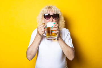 Blond old woman in a white T-shirt with sunglasses holding a glass of beer on a yellow background