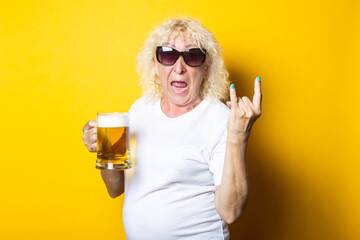 Surprised blonde old woman holding a glass of beer and showing a rocker goat on a yellow background