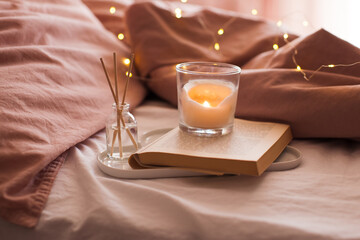 Burning candle with aroma sticks in bottle on tray with open book in bed over glowing Christmas lights close up. Cozy atmosphere at home. Good morning. Selective focus.