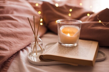 Burning candle with aroma sticks in bottle on tray with open book in bed over glowing Christmas lights close up. Cozy atmosphere at home. Good morning. Selective focus.