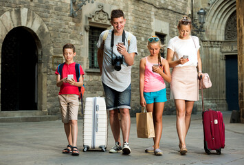 Smiling young family of four with suitcases walking along city using phones