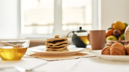 A stack of sweet tasty pancakes, honey in a bowl, teapot, croissants and various fruits served for breakfast on the table