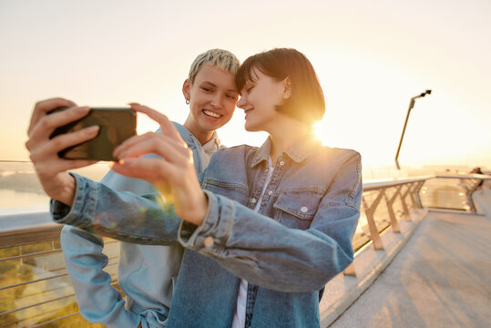 Lesbian couple standing on the bridge, smiling while taking a selfie picture, watching the sunrise together