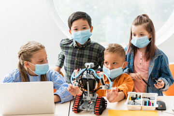 Selective focus of multiethnic classmates in medical masks constructing robot near laptop in classroom