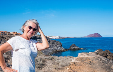Fototapeta na wymiar Attractive senior woman white haired standing on the cliffs enjoying vacation or excursion and freedom in seascape. Active retired elderly people