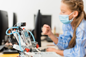 Selective focus of robot on table near schoolgirl in medical mask in classroom