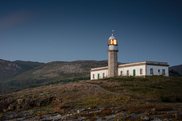 Fototapeta na wymiar Lariño lighthouse at sunset with the light on and under blue sky. Carnota, Galicia, Spain