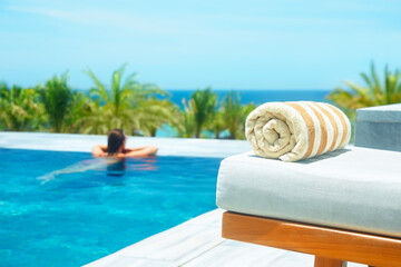 Rolled up towel on lounge chair by luxury outdoor pool to background of palm trees and ocean. Focus on foreground