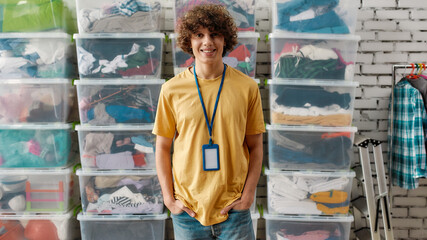 Caucasian guy smiling at camera, posing in front of rack and boxes full of clothes, Young volunteer working for a charity, donating apparel to needy people