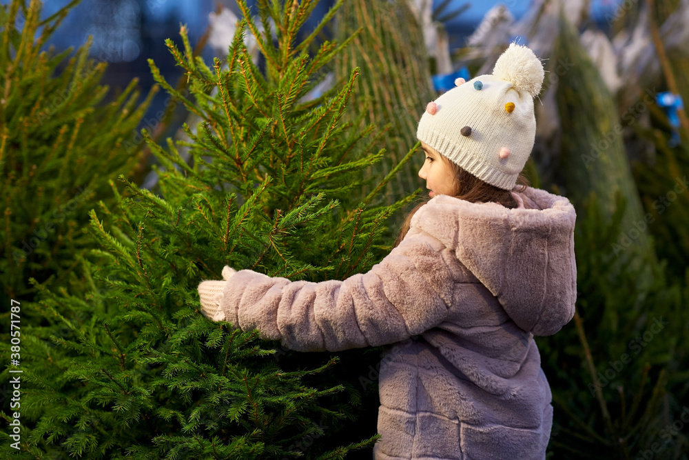 Poster winter holidays and people concept - happy smiling little girl choosing christmas tree at street market in evening