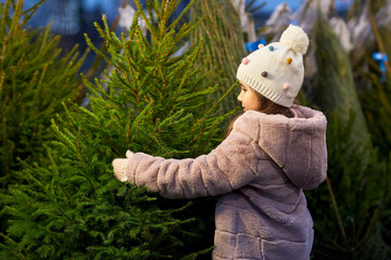 winter holidays and people concept - happy smiling little girl choosing christmas tree at street market in evening