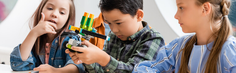 Panoramic shot of multicultural schoolkids looking at robot in stem school