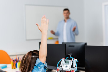 Selective focus of schoolgirl with raised hand near robot and computers in stem school