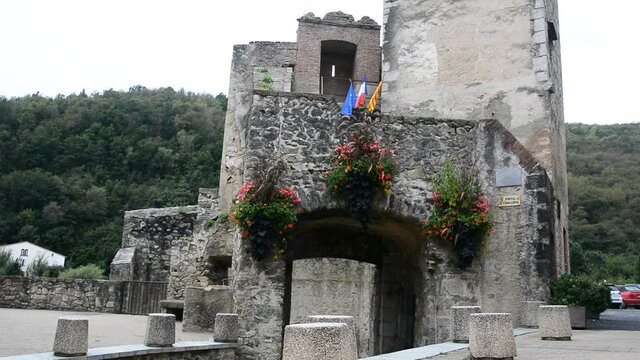 Still image of the entrance to the fortress within the medieval village of Prats de Mollo la Preste, in it you can see several red flowers and the Catalan, French and European flags