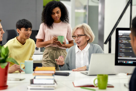Aged woman, senior intern chatting with her young colleagues, Friendly workers talking with new employee during coffee break at work