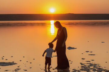 Mother and son at sunset, silhouettes against the sunset. Mom walking and having fun with her kid on the beach at sunset