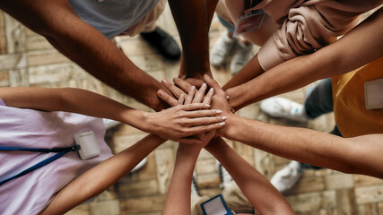 Top view of diverse young volunteers putting their hands on top of each other like a real team while standing in charitable organization office - Powered by Adobe