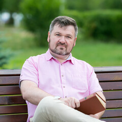 Fototapeta premium Adult Caucasian man 40-45 years old sits on a bench with a book in his hand.