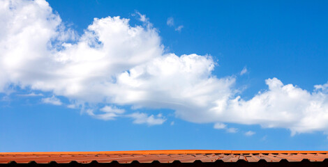 A large beautiful long white cumulus cloud in a blue clear sky over a brown metal roof.