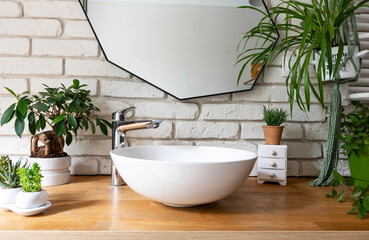 Modern interior with mirror on white brick wall and stylish sink and faucet in industrial bathroom. Bright bath  with green plants and wooden furniture. 