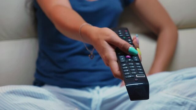 Close up of switching tv programs sitting on sofa using remote control. Shot of controller in hands of person pointing to TV pressing the button and changing channels sitting in front of television