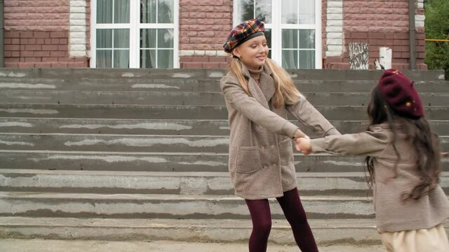 Cute happy girls holding hands and rotating near stairs. Adorable cheerful sisters in trendy coats and berets having fun together near steps and building
