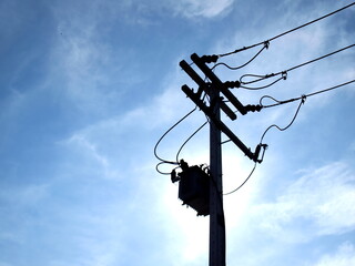 Silhouette of a transformer on a pole. Complete with fuse and an electric cable for converting high voltage AC to low voltage. On the blue sky background there are white clouds. Selective focus