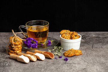 Decoration of hot english tea in luxury glass with saucer with multi grain cookies stack and in white ceramic bowl on gray table.