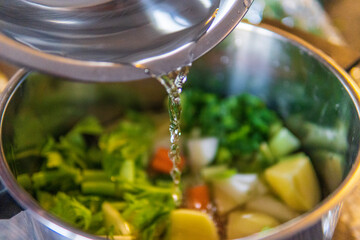 Cooking vegetables soup / stock: adding water to fresh uncooked ingredients of vegetables and root veggies in a pot. Potato, onions carrots, celery, leek, coriander, parsley and dill. Selective focus
