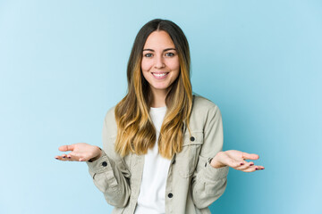 Young caucasian woman isolated on blue background makes scale with arms, feels happy and confident.