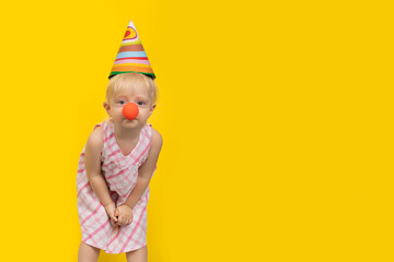 Cute fair-haired girl in festive hat and clown nose looking at camera. Holiday or birthday party concept. Copy space.