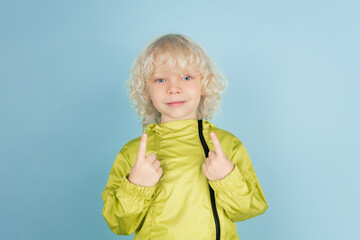 Pointing up. Portrait of beautiful caucasian little boy isolated on blue studio background. Blonde curly male model. Concept of facial expression, human emotions, childhood, ad, sales.
