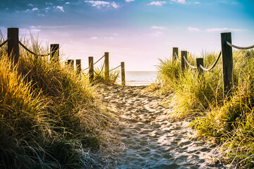 Long shadows fall over sandy pathway with grass reeds and wooden posts on each side leading to a beautiful sea bay