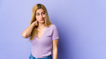 Young blonde woman isolated on purple background touching back of head, thinking and making a choice.