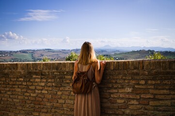 A young blonde woman is looking at the view beyond a brick wall (Corinaldo, Marche, Italy, Europe)