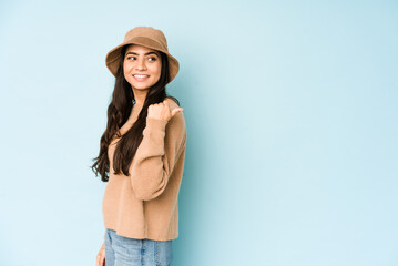 Young indian woman wearing a hat isolated on blue background points with thumb finger away, laughing and carefree.