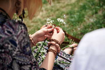 Close-up picture of woman, wearing colorful bracelets, making flower camomile wreath on green field on sunny summer day. Eco tourism in rural countryside. Young festival handmade activity.