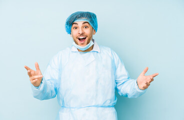 Young arabian surgeon man isolated against on a blue background receiving a pleasant surprise, excited and raising hands.