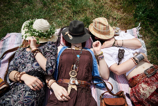 Three hippie women, wearing colorful boho style clothes and straw hats, lying, relaxing on green grass of field on sunny summer day. Eco tourism in rural countryside. Friends traveling together.