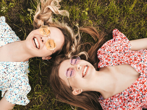 Two young beautiful smiling hipster girls in trendy summer sundress.Sexy carefree women lying on the green grass in sunglasses.Positive models having fun.Top view