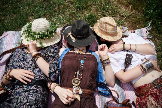 Three hippie women, wearing colorful boho style clothes and straw hats, lying, relaxing on green grass of field on sunny summer day. Eco tourism in rural countryside. Friends traveling together.