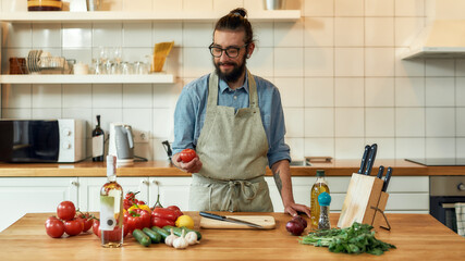 Obraz premium Young man, Italian cook in apron holding, looking at tomato ready for preparing healthy meal with vegetables in the kitchen. Cooking at home concept