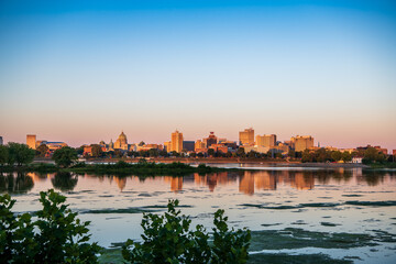 A photo of Riverfront Park at sunset seen from across the Susquehanna River at sunset. The buildings reflections are shown in the river. 