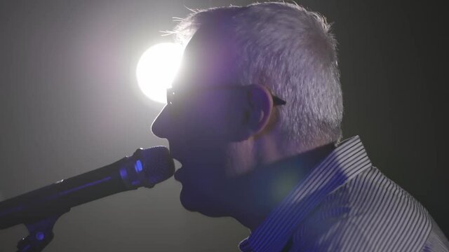Man singing in dark and blue lighting, side angle handheld close up while backlit.