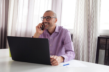 Mature attractive moroccan entrepreneur bearded man wearing shirt sitting on a white desk table with coffee and laptop working from home happy and confortable while makes phonecall and drinks coffee.