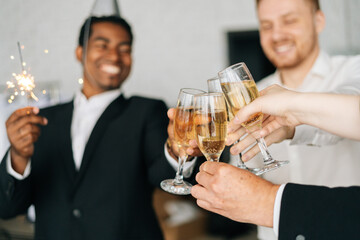 Close-up view of business team of multinational employees holding Bengal lights, clinking glasses of champagne and smiling cheerfully at corporate New Year or Happy Christmas eve.