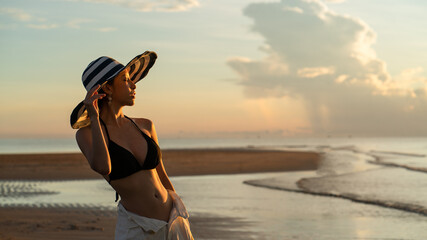 Woman in top bikini and white long pant wearing hat on the beach with a beautiful sunrise or sunset in background.