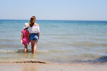 Back view mother and little daughter holding hands standing in sea water looking into horizon, copy space for text. Family travel and summer vacation concept